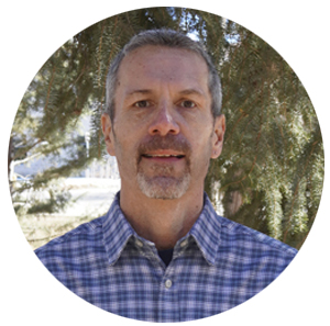 man in blue plaid shirt stands in a wooded area