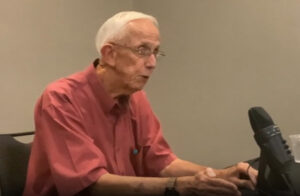man in red shirt and white hair sits in front of a microphone.
